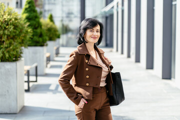 Purposeful middle age caucasian brunette woman in brown suit standing outdoor on summer shiny day. Cute smiling Italian pretty woman.