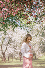 A beautiful pregnant woman walks in a blooming garden. Delicate pink peachy silk dress and white cardigan, long blond hair. Expecting a child, fertility, harmony