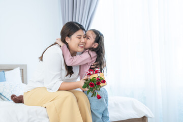 Obraz na płótnie Canvas Cute mixed race child daughter, Caucasian and Asian, kissing and giving gift box and roses flowers to young mother for mother's day or birthday in bed at home with love