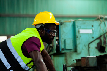 A black man African American worker wearing glasses in a yellow hard hat works machinery in an industrial factory.
