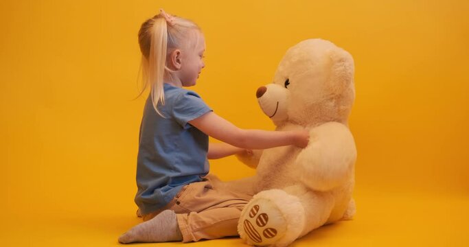 Preschooler fair-haired girl playing with big teddy bear. Child sits on the floor with soft bear toy. Yellow background