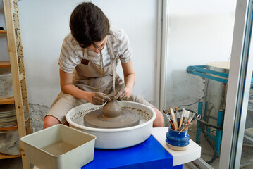 young ceramist on the lathe or potter's wheel working and shaping clay inside a pottery studio, creating creative shapes with tools. natural light