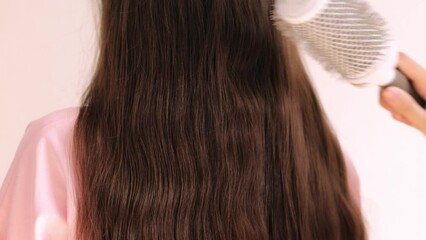 Combing light brown hair with a white comb on an isolated pink background. A young girl in a pink T-shirt in a beauty salon. Rear view of the client's back and the master's hands.