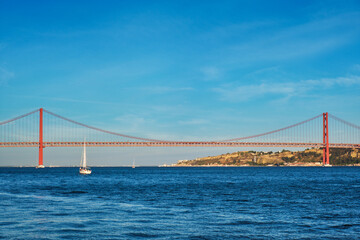 Fototapeta na wymiar View of 25 de Abril Bridge famous tourist landmark over Tagus river and a tourist yacht boat at sunset. Lisbon, Portugal