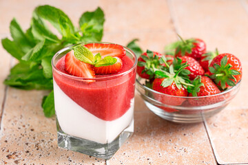 Panna cotta sweet homemade Italian dessert of cream thickened with gelatin with fresh strawberry sauce decorated with berries and mint leaf served in glass on kitchen table with ingredients in bowl