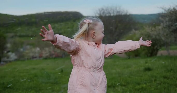 Happy little girl with blonde hair and two ponytails in pink jacket stands in green meadow with her hands raised.
