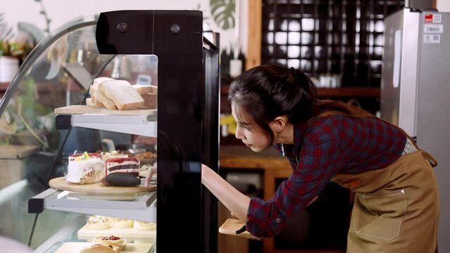 Cafe worker is opening bakery cabinet, bent down use tongs pick cake in refrigerator out put them on sall tray, that is served customers who order eat in store, when finished, close cabinet as before.