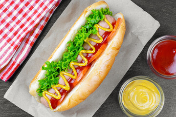 Homemade Hot Dog with yellow mustard, ketchup, tomato and fresh salad leaves on black slate background. Fast food, street food, american cuisine