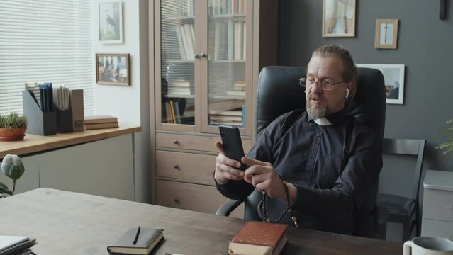 Mature Caucasian priest wearing eyeglasses and earbuds sitting at wooden desk in his office talking on video call on smartphone
