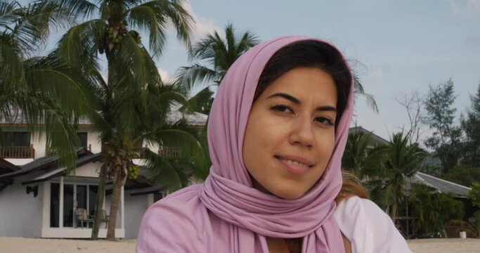 A Smiling Muslim Travel Blogger Poses Against A Scenic Backdrop Of A Beach And Vacation Bungalows. Her Vibrant Attire And Joyous Expression Reflect Her Passion For Exploration.