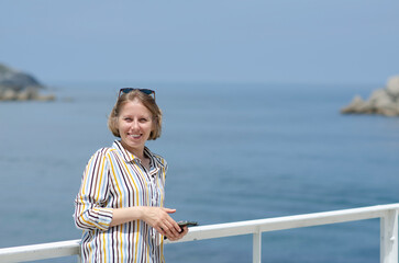 Beautiful caucasian blonde woman with square haircut and sunglasses, in striped shirt is smiling and holding a phone in her hand. Concept of freedom and travel in summer. Blogging and working online.