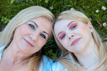 Close-up portrait of faces of two blonde mom and daughter, similar to each other against background of green grass.