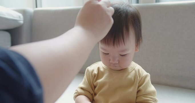 Happy Asian family mother tenderly comb hair make hairstyle of her baby, little daughter child toddler girl sits on sofa and mother combing hair of kid using comb daily morning beauty procedures