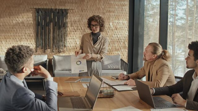 Full zoom in shot of young Caucasian woman in casual clothes standing in corporate meeting room in office on bright sunny day, holding document with pie chart, and talking to diverse male colleagues