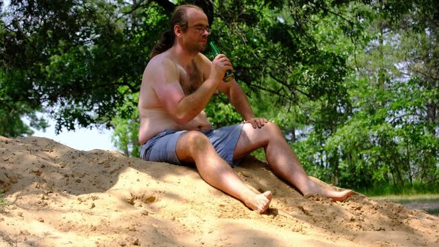 Barefoot and shirtless young man sitting in blue shorts on top of pile of sand holding and drinking beer while taking short brake from work.