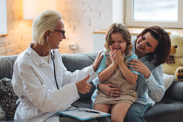 Kind senior female paediatrician doctor examining little child sitting on mother's laps, during home visit or clinic check up. Concept healthcare medical assistance, insurance. Prescribing treatment