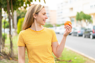 Young blonde woman holding an orange at outdoors with happy expression