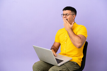 Young man sitting on a chair with laptop yawning and covering wide open mouth with hand