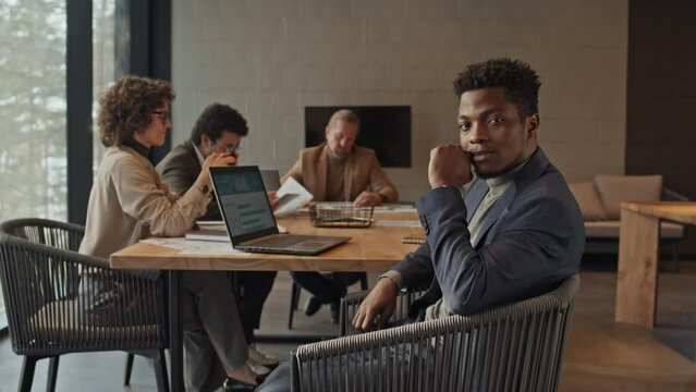 Medium portrait shot of young African American businessman in suit and turtleneck sitting in chair in meeting room, posing for camera, while multiethnic startup team is working together in background