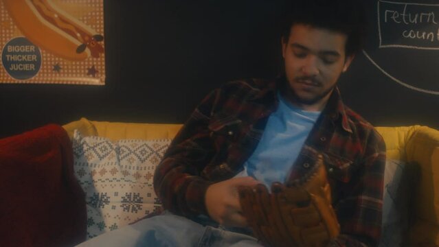 Young Middle Eastern man sitting relaxed on sofa wearing baseball glove playing with ball, 90s aesthetics