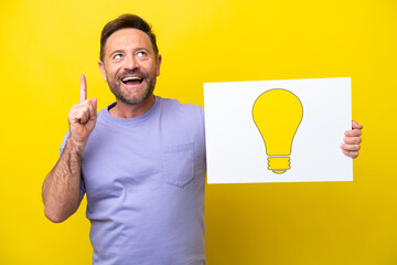 Middle age caucasian man isolated on yellow background holding a placard with bulb icon and thinking
