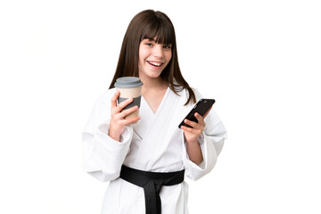 Fototapeta premium Little Caucasian girl doing karate over isolated background holding coffee to take away and a mobile
