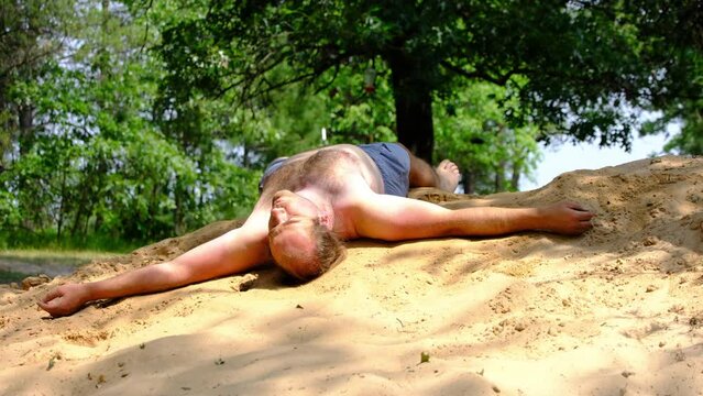 Young shirtless and barefoot male wearing blue shorts waking up and gets disoriented while falling asleep on top of pile of sand after back yard pool party.