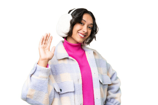 Young Argentinian woman wearing winter muffs over isolated background saluting with hand with happy expression