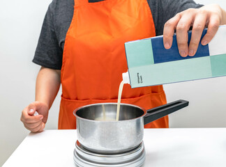 Professional chef with orange apron pouring milk from container into stainless steel pot. Person pouring whipped cream into saucepan on white table, preparing a recipe for homemade bakery and pastry.