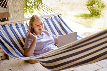 A young girl lies in a hammock with a laptop and communicates through a video call. 