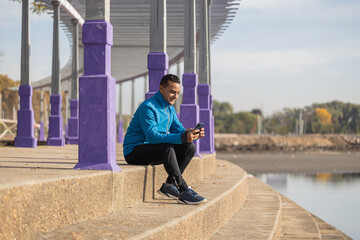 Young latino man in sportswear looks at his mobile phone sitting on some stairs in a public park with copy space.