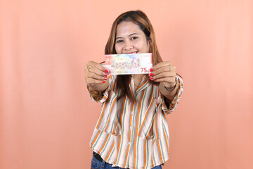 Portrait of happy Indonesian Muslim woman holding rupiah money isolated over peach background. 