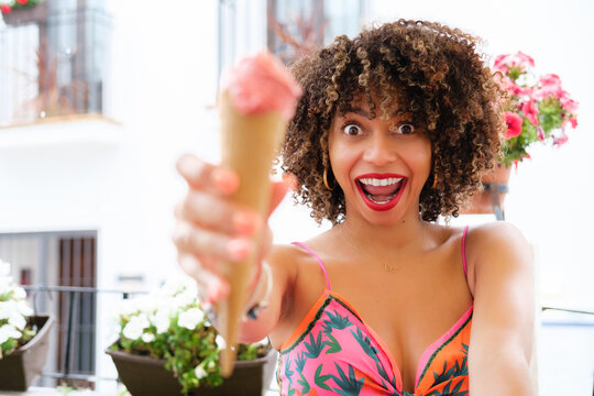 A girl with afro hair and dressed in bright colors displays, extremely cheerful, a strawberry ice cream cone.
