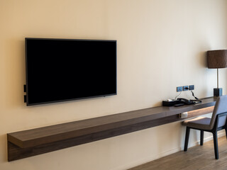 Mockup blank space on smart TV black screen over the empty long brown walnut wooden table shelf with chair at working area on white wall background with yellow warm lighting in the hotel bedroom.