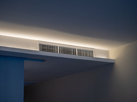 Air conditioning wall mounted ventilation system on ceiling in the white hotel room with dim light. Hotel room air ventilation grill on the wall.
