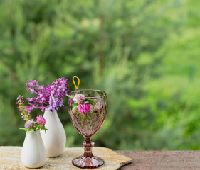 A glass with a drink and flowers in a white vase on the terrace against the backdrop of a green garden