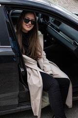 beautiful young girl with brown hair green eyes in the bright trench sits in a black car