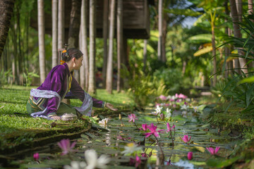 A pretty young Asian woman wearing an elegant luxurious traditional Chiang Mai dress costume sits on a natural green lawn by a stream with a beautiful pink lotus flower nearby.