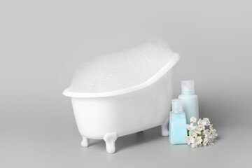Obraz na płótnie Canvas Small bathtub with foam, bottles of cosmetic products and flowers on light background