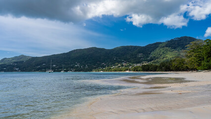 The waves of the turquoise ocean spread over the beach. Wet sand glistens. Yachts in the distance. A green hill against a background of blue sky and clouds. Seychelles. Mahe. Beau Vallon.