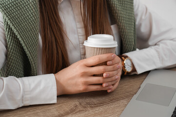 Woman holding takeaway cup of coffee at table with modern laptop