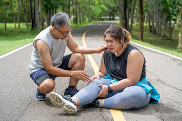 Overweight young woman with injured knee sitting on the running track at local park with both hands grabbing on her trouble knee while a middle age man kneeling beside her