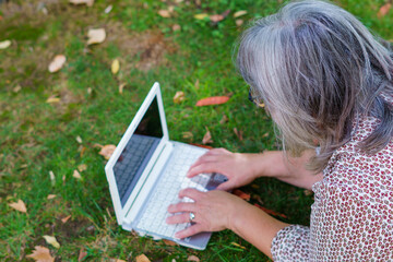 top view of a white-haired woman working on her laptop computer lying on the grass in a park.