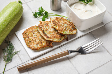 Board of tasty zucchini fritters with sour cream on white tile background