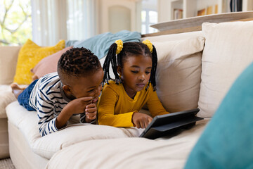 African american siblings using digital tablet while lying and relaxing on sofa in living room