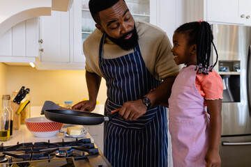 African american father with smiling daughter cooking pancakes in pan in kitchen