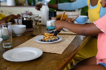Fototapeta na wymiar Midsection of african american girl eating pancakes with fork and table knife at dining table