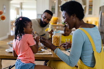 African american mother feeding pancake to daughter while having breakfast with family at table