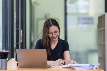 Fototapeta premium A beautiful young Asian businesswoman is saving and checking work and documents with her laptop computer at her desk with a bright smile on her face.