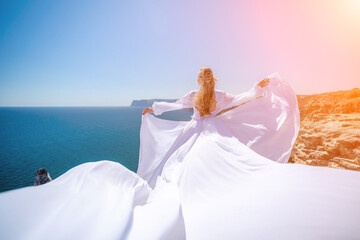 Fototapeta na wymiar woman sea white dress. Blonde with long hair on a sunny seashore in a white flowing dress, rear view, silk fabric waving in the wind. Against the backdrop of the blue sky and mountains on the seashore